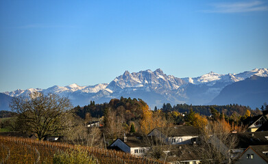 View of the Glarner Alps with Mount Glarnisch covered in snow, seen above a Swiss village surrounded by autumn trees under a clear blue sky