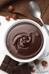 Melted chocolate in bowl, pieces, nuts and spoon on white marble table, flat lay