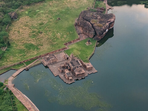 Aerial view of Bhutanatha Temple complex reflecting in the still, dark waters of the lake, framed by rugged cliffs and verdant greenery, Badami, Karnataka, India.