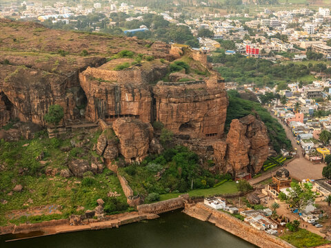 Aerial view of the rugged, ochre-toned Badami cave temples carved into the sandstone cliffs, overlooking the serene Agastya Lake, Badami, Karnataka, India.