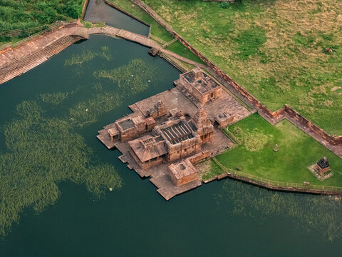 Aerial view of Bhutanatha Temple complex reflecting in the still, dark waters of the lake, framed by rugged cliffs and verdant greenery, Badami, Karnataka, India.
