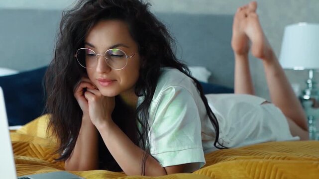 Caucasian sexy girl with long dark hair wearing glasses is lying on bed and watching movie on her computer. Beautiful woman relaxing in her bed, smiling, looking with interest and moving her legs.