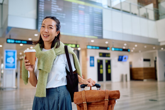 Happy asian woman traveler enjoying a cup of coffee while waiting for her flight at the departures gate inside an airport