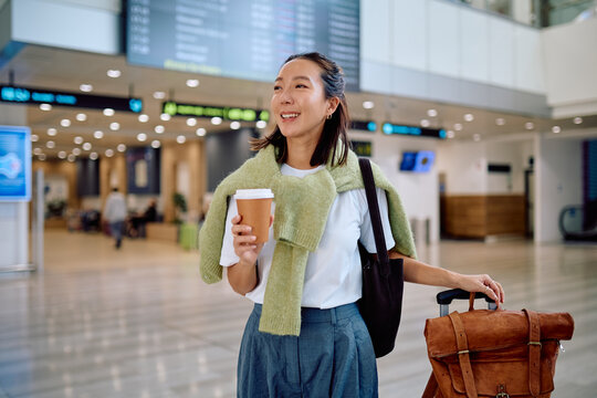 Young Asian woman holding a coffee cup, pulling luggage, and smiling while walking in a bright airport terminal for departure