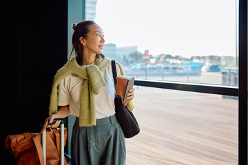 Young woman standing with luggage, holding coffee and phone, looking out window while waiting in an airport terminal © we.bond.creations