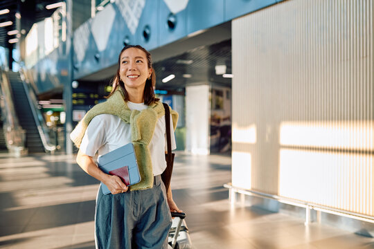 Smiling woman with passport and tablet pulling luggage, arriving or departing from modern airport terminal