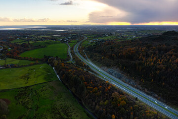 Aerial view of the M1 motorway with cars surrounded by autumn forest and green fields during a cloudy evening in rural Ireland