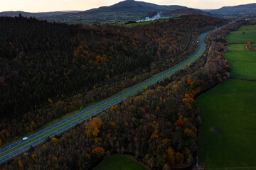 Aerial view of the M1 motorway with cars surrounded by autumn forest and green fields during a cloudy evening in rural Ireland