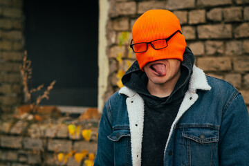 A young man stands in front of a brick wall wearing an orange beanie and glasses playfully sticking...