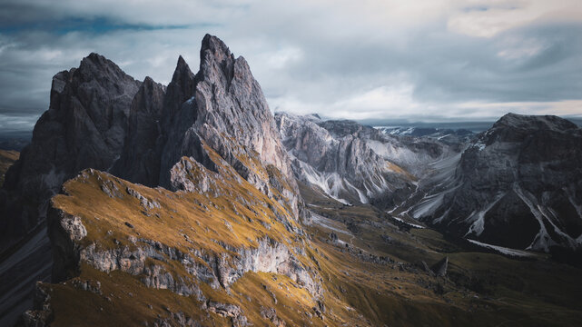 Aerial view of jagged peaks pierce the sky, a symphony of gray stone and golden slopes under a watchful, cloudy expanse, Dolomites, Italy.