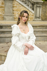 Obraz premium figure of female blonde model wearing white fantasy wedding gown. romantic sitting pose on staircase of historical fairytale castle location 