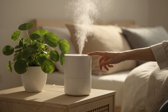 A woman's hand turns on a white ultrasonic humidifier on a nightstand in a bedroom