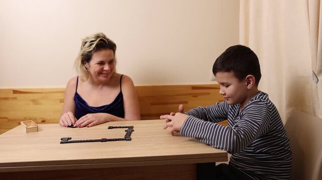 A woman and a boy are playing dominoes while sitting at a table.