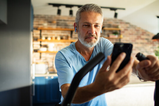 Mature man focused on indoor cycling with smartphone at home - Powered by Adobe