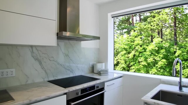 Modern kitchen with sleek white cabinets, marble accents, stainless appliances, and a window overlooking trees