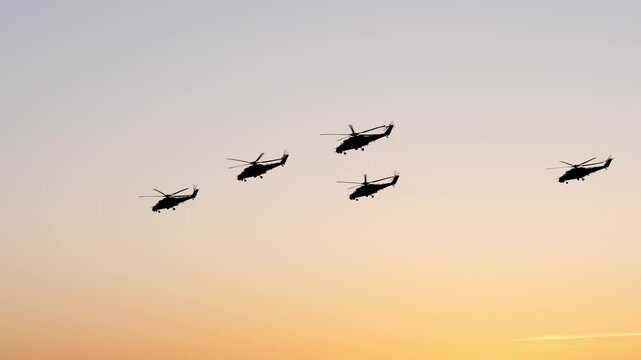 Multiple military helicopter silhouettes flying together in formation during a mission or airshow parade. Against a beautiful orange and blue sunset sky, symbolizing power warfare and precision