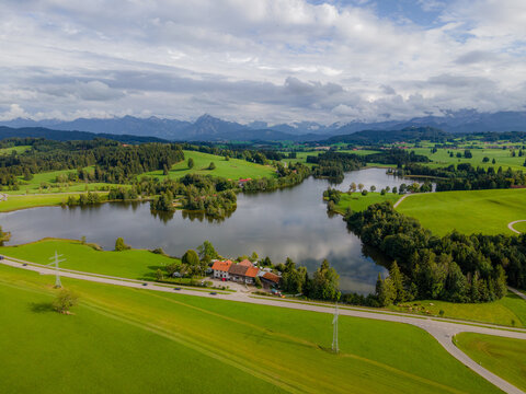 Aerial view of the serene lake mirroring the cloudy sky, bordered by lush green fields and shadowed forests, framed by distant, majestic mountains, Ruckholz, Bayern, Germany.