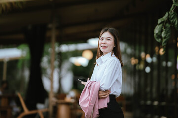 A young woman in business attire smiles while using her smartphone outdoors, holding a pink blazer and appearing engaged and cheerful.