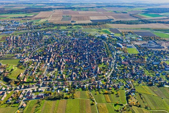 Aerial view of a medieval town surrounded by patchwork fields and vineyards, a tapestry of russet roofs and emerald pastures blending seamlessly, Rouffach, Grand Est, France.