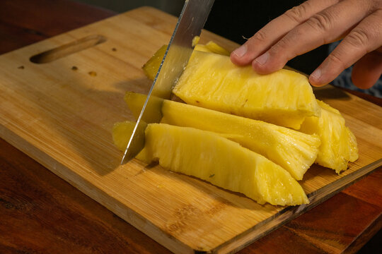 A freshly peeled pineapple is being sliced into chunks on a wooden cutting board