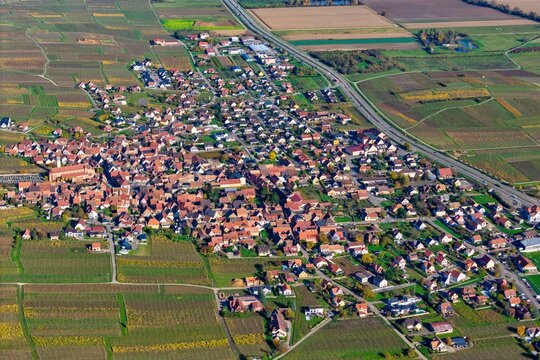 Aerial view of terracotta rooftops nestled amidst a patchwork of vineyards, a symphony of autumnal hues blanketing the landscape, Rouffach, Grand Est, France.