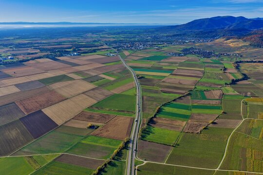 Aerial view of patchwork fields, a ribbon of road cutting through the landscape towards distant mountains under a clear sky, Rouffach, Grand Est, France.