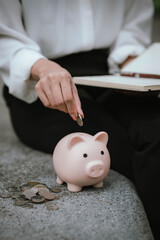 A woman wearing a white shirt is saving money by putting a coin into a pink piggy bank, symbolizing financial planning and saving for the future.