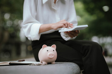 A person saves money by putting coins into a pink piggy bank while holding a calculator, symbolizing financial planning, saving, and budgeting.