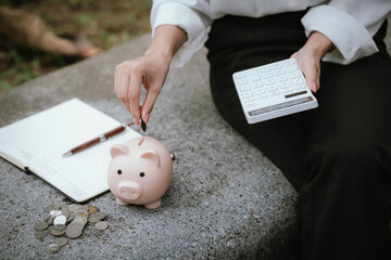A person saves money by putting coins into a pink piggy bank while holding a calculator, symbolizing financial planning, saving, and budgeting.