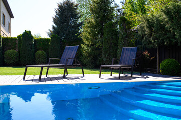 Two wicker sun loungers on a wooden deck near a clear blue pool in a private yard, surrounded by lush decorative trees and green lawn. Bright sunny day