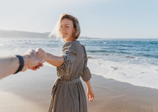 Beautiful woman leads partner by hand on the ocean. Happy couple on vacation together. Couple holding hands and running on beach. Follow me - concept of travel photo. Beach Holidays.