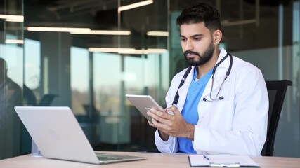 Confident doctor in white coat using digital tablet while sitting at workplace in a modern hospital office. Focused handsome medical worker physician chats online with patient or works in application - Powered by Adobe