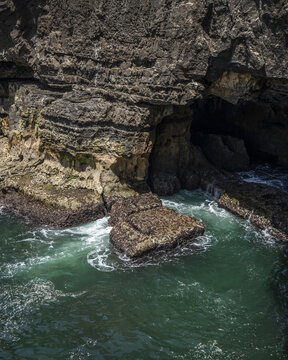 View of the rugged coastline where dark, jagged cliffs meet the churning emerald sea, creating a dramatic interplay of shadow and light, Cascais, Lisbon, Portugal.
