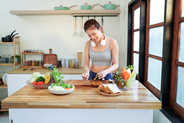 Healthy weight loss nutrition. Slim lady in sportswear cooking fresh vegetable salad, mixing ingredients standing at kitchen table at home