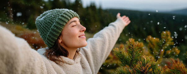 A woman in a cozy jacket and knitted hat standing with raised hands in nature during sunset. Blurred winter forest in background.