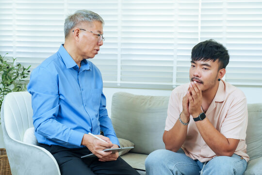 Asian adult man sitting with clasped hands while discussing feelings with senior psychiatrist in therapy session, showing reflection and openness during professional psychological counseling