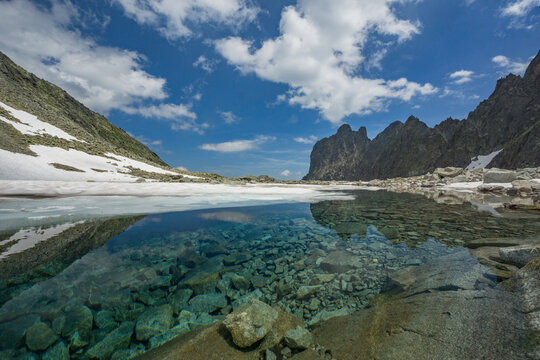 View of crystal waters reflect towering peaks under a vast sky, framed by snow-dusted slopes and rugged rocks, Zamrznute pleso tarn, Presovsky kraj, Slovakia.