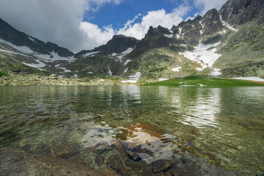 View of crystal-clear waters reflecting jagged peaks dusted with snow under a sky brushed with clouds at Zamrznute pleso tarn, Tatranska Javorina, Presovsky kraj, Slovakia.