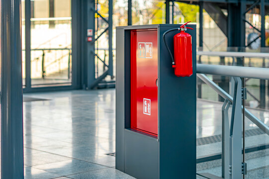 Red fire hydrant cabinet and mounted extinguisher on metal support structure inside modern public building corridor with safety signage and emergency equipment