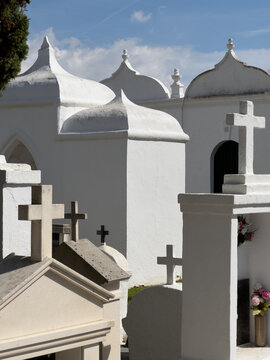 Panteones y cruces blancas en cementerio mediterr&aacute;neo bajo cielo azul con nubes