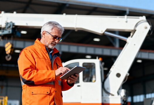 Mature engineer using a digital tablet at an industrial site. Construction manager working with a crane in the background. Modern technology in heavy industry
