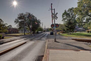 View of the sun shining above a railroad crossing with a long road ahead, trees lining the sides, and cars parked along the street, Loveland, Ohio, United States.