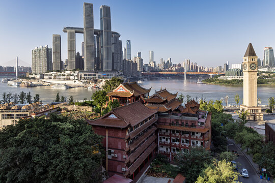 View of Thousand Buddha Temple against towering modern skyscrapers and a clock tower, all overlooking a wide river in Chongqing, China.