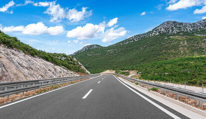 Scenic mountain highway curving through green hills under a bright blue sky with fluffy white clouds on a sunny summer day.