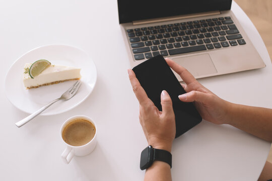 Close-up of hands using smartphone at desk with laptop, espresso and lime cheesecake, reflecting digital lifestyle, mobile productivity and remote work during a casual coffee break