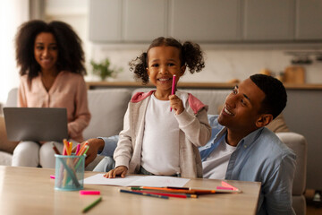 African American dad drawing with little daughter, spending quality time babysitting with child while mother working on laptop, sitting nearby