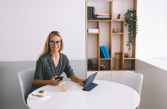 Confident young freelancer using smartphone and tablet while smiling at camera during remote work session, embracing multitasking and mobile tech in a digital lifestyle - Powered by Adobe