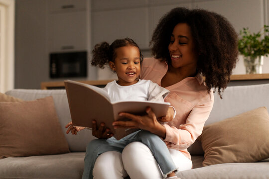 Black mother reading book to little cute daughter sitting together on sofa, enjoy good life hobby and pastime at home. Parenthood, upbringing, children development concept