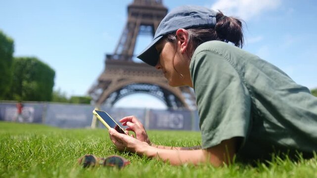 Person in cap and casual clothes lies on lush grass near sunglasses, holding smartphone with blurred eiffel tower in background, enjoying travel, technology and sunny park leisure