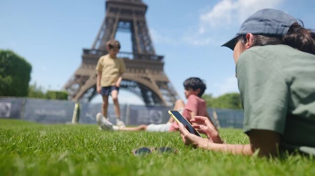 Person in cap and casual clothes lies on lush grass near sunglasses, holding smartphone with blurred eiffel tower in background, enjoying travel, technology and sunny park leisure
